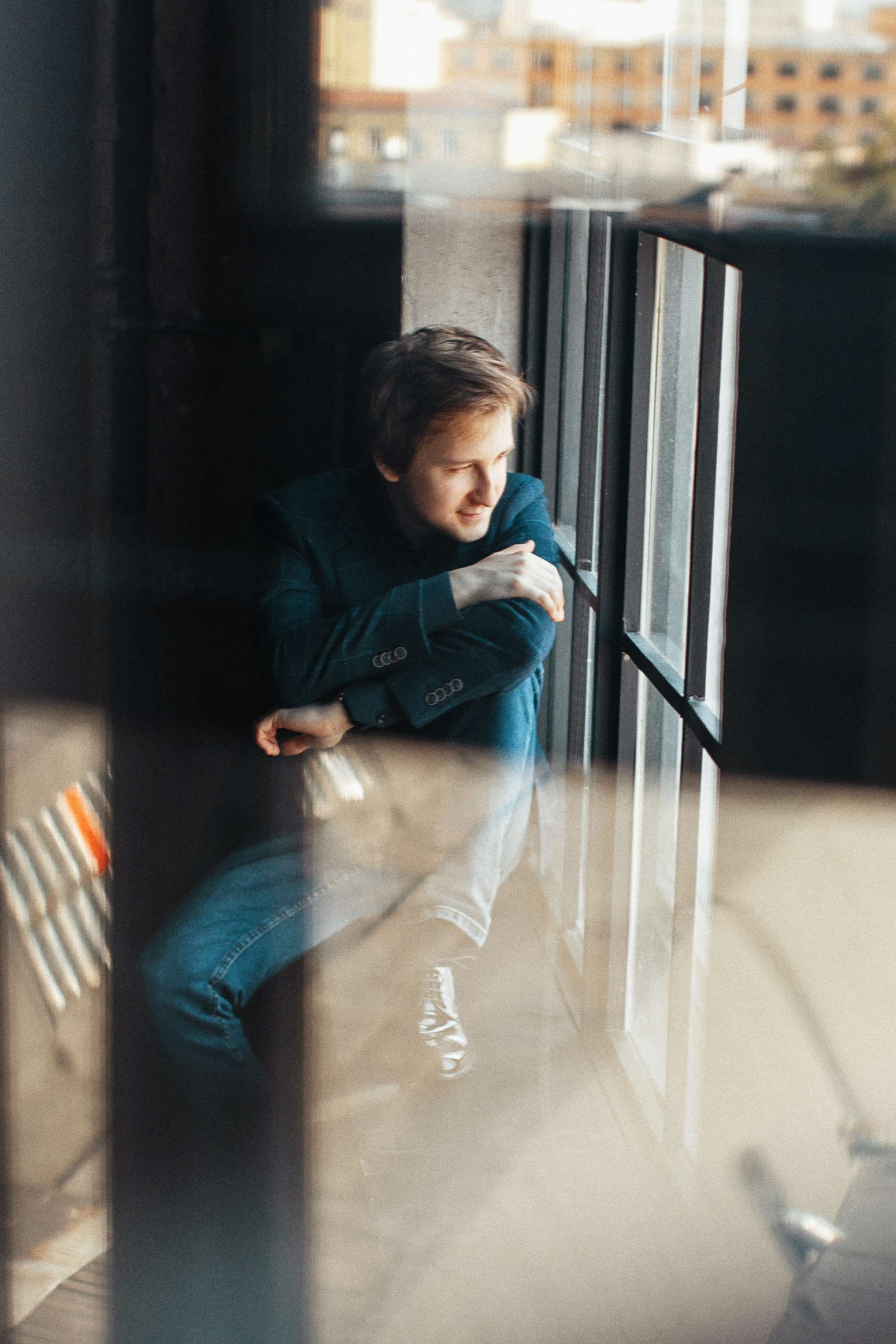 Dmitry Masleev pose pour un portrait de trois quart. Un jeu de reflet montre des tours d'immeubles au loin. 