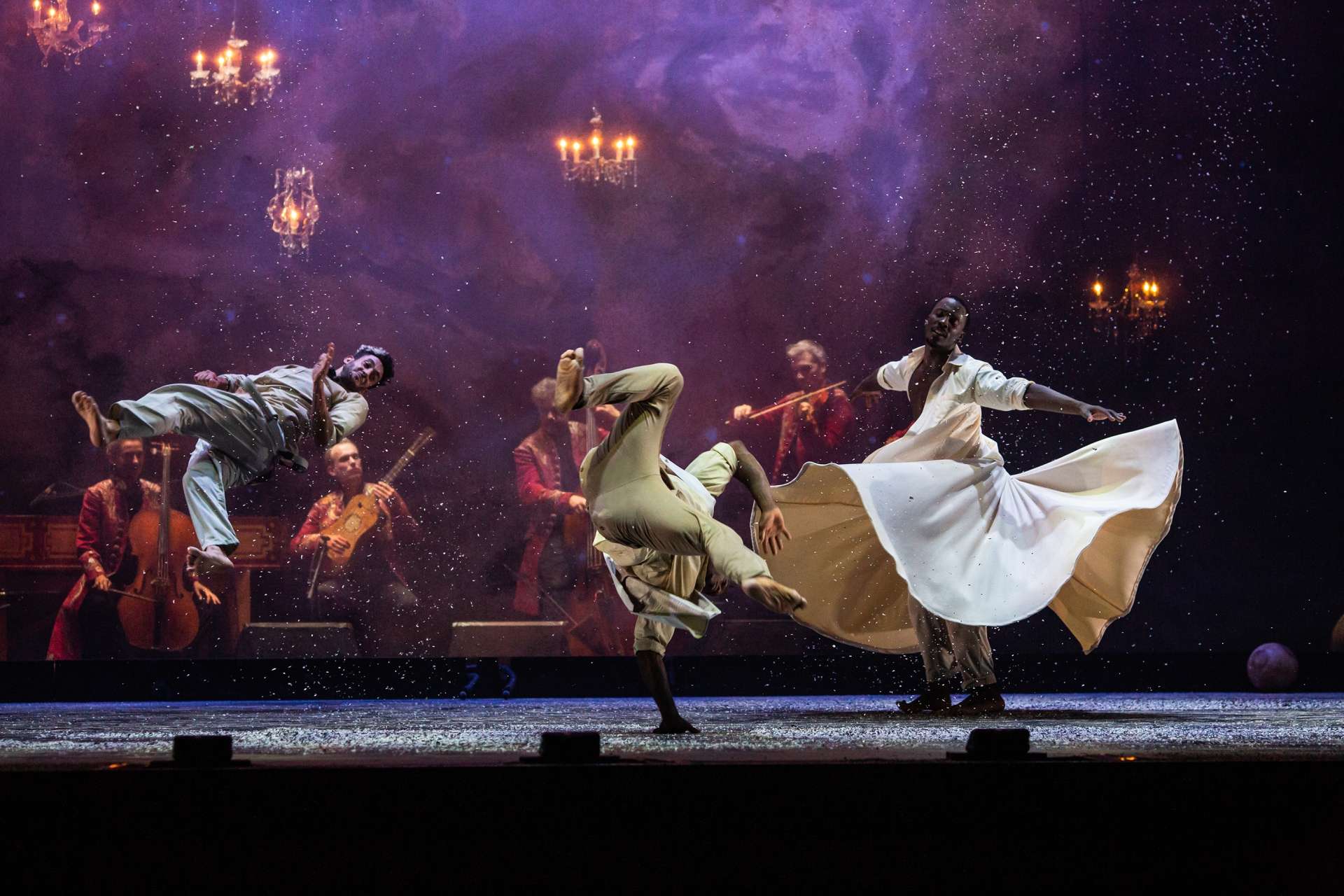 Féerie du spectacle Folia avec deux danseurs hip-hop et un derviche tourneur en bord de scène et en fond de scène musiciens et musiciennes. Atmosphère féerique et voluptueuse avec une lumière chaleureuse et des lustres chandeliers. 