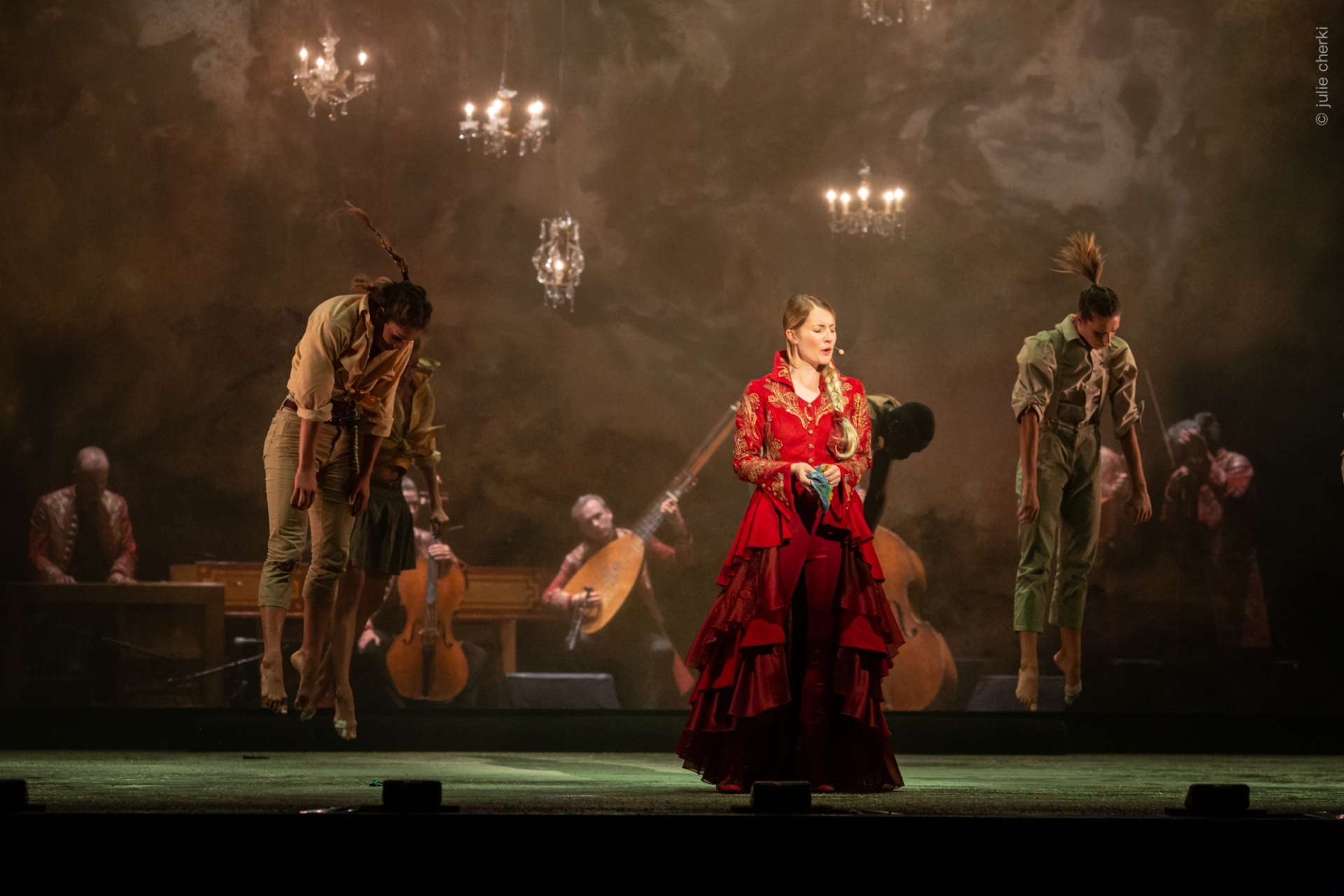 Une chanteuse lyrique illumine la scène avec sa majestueuse robe rouge. De chaque côté, deux danseuses l'entourent et les musiciens jouent toujours en fond de scène. Les chandeliers illuminent aussi la scène. 