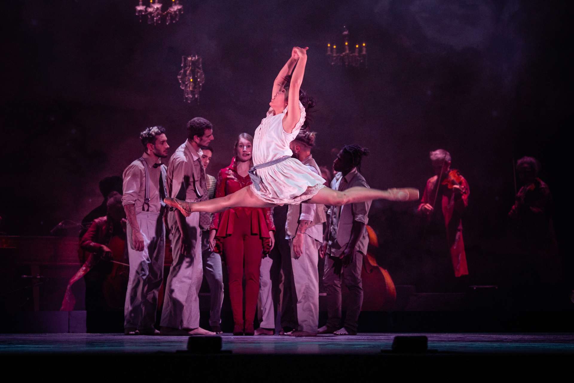 Au premier plan, une danseuse en pointes fait un grand écart sauté. En fond de scène musiciens et chanteurs se regroupent. 