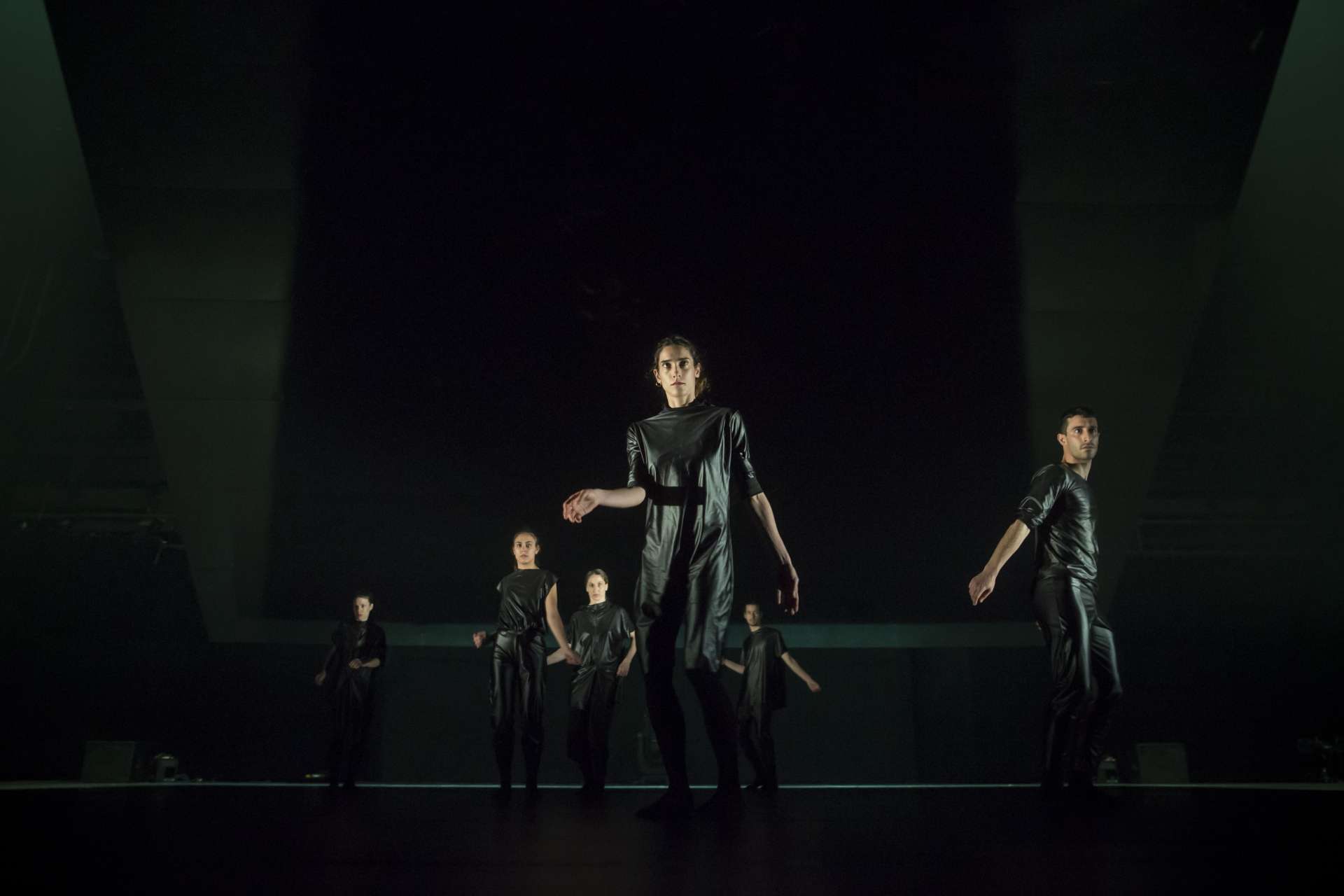 Sur scène fond noir, cinq danseurs et danseuses vêtues de noir effectuent une chorégraphie. 