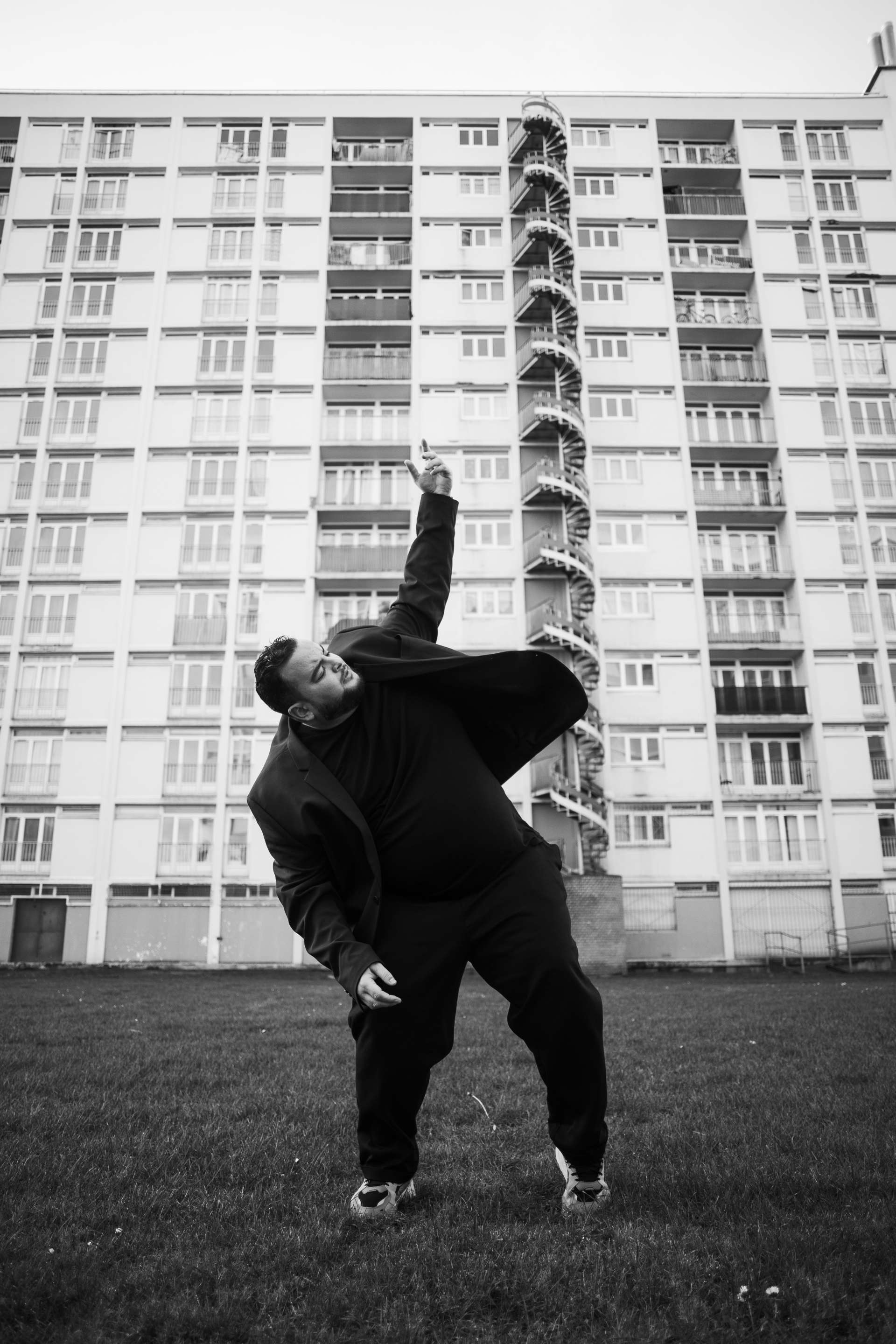 Le danseur et chorégraphe Sofiane Chalal danse en costume noir devant une grande tour d'immeuble. 