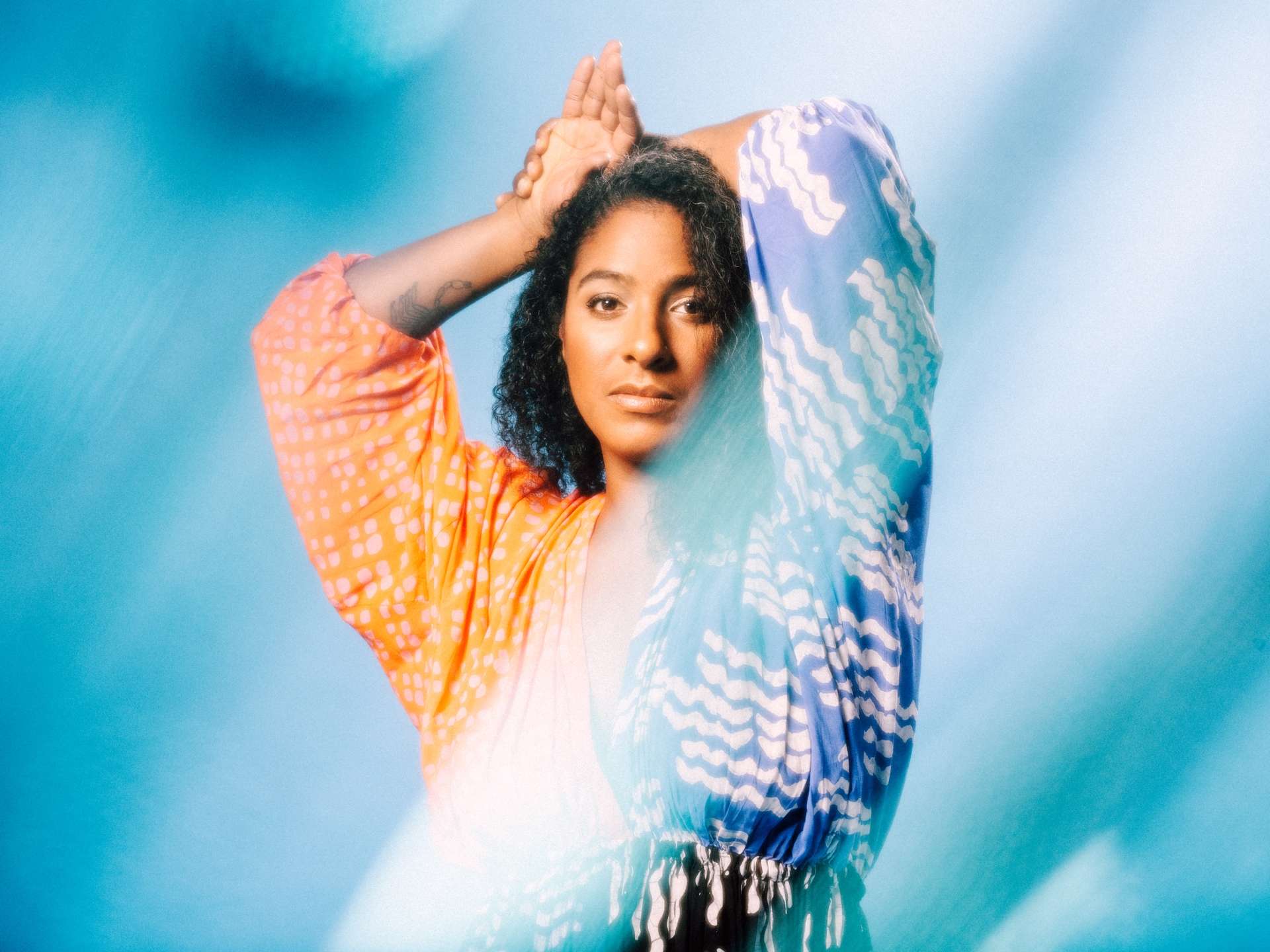 Portrait posé de l'artiste Leyla McCalla sur fond bleu et vêtue d'une tenue fluide et colorée. 