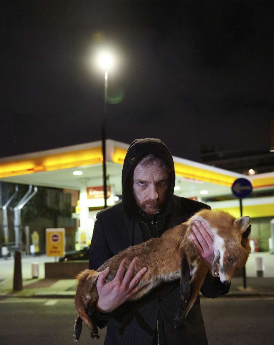 Photo de nuit. Homme avec capuche au premier plan tenant un cadavre de renard dans les bras. Station service en arrière plan
