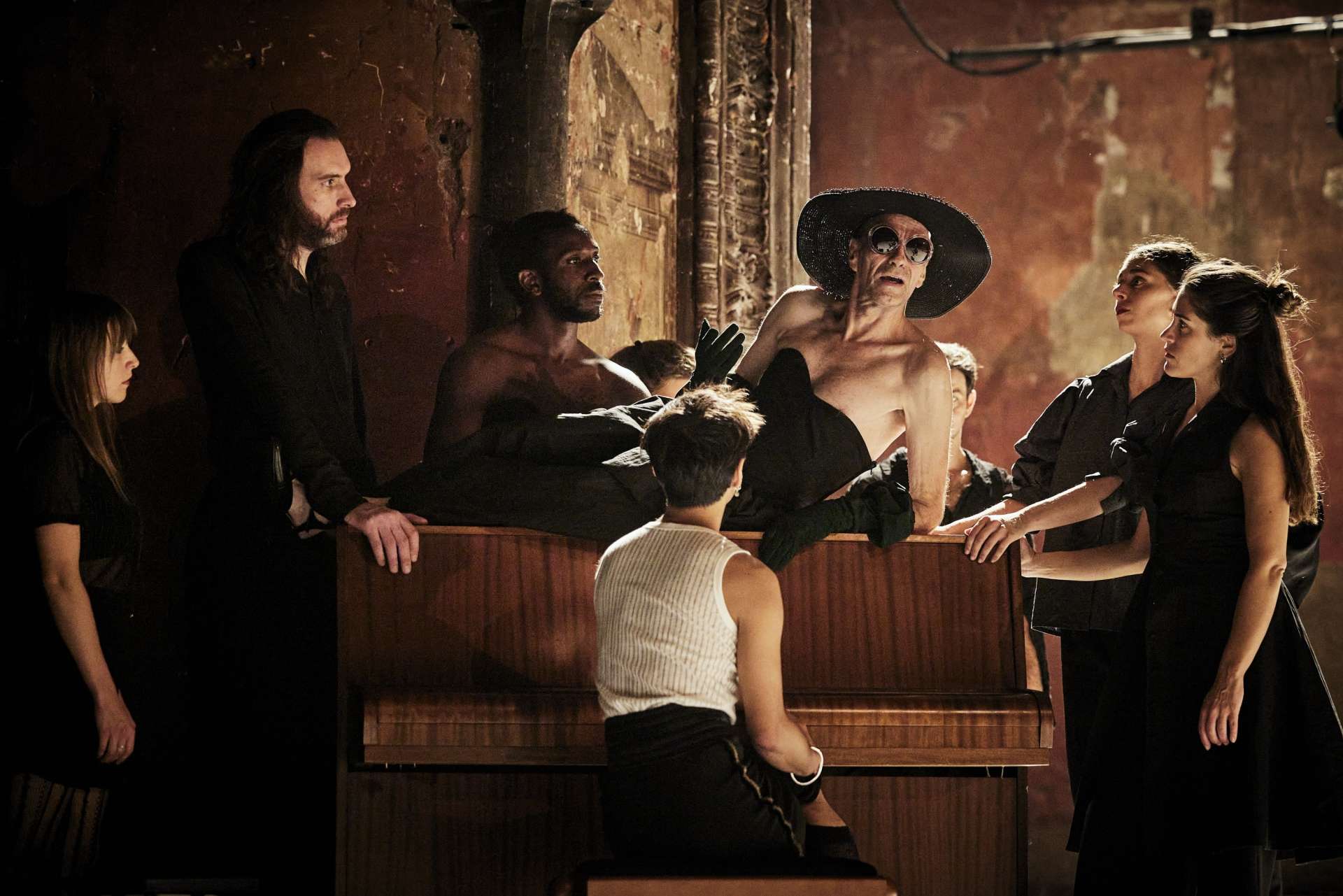 Ambiance cinéma. Une homme habillé en femme, vêtu d'une robe noire bustier, d'un grand chapeau et de lunettes de soleil est allongé sur un piano marron. Autour de lui, hommes et femmes, en noir aussi, sont debout, accoudés ou assis et le regarde. 
