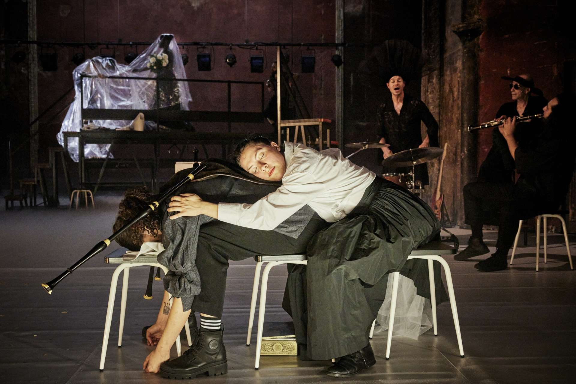 Un homme est assis et plié en deux. Une femme est assise derrière et allongée sur son dos. Au fond, des musiciens. 