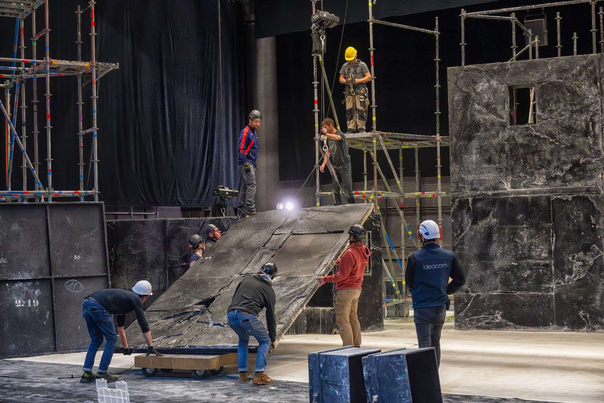 Des techniciens lèvent un décor en plâtre noir grâce à un moteur pour l'accrocher à un échafaudage.