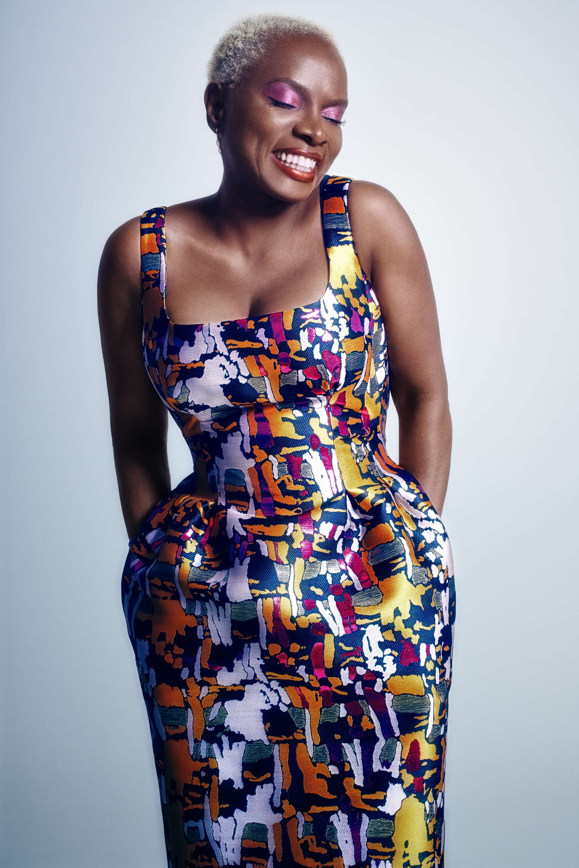 La souriante diva Angélique Kidjo pose sur un fond gris avec une robe colorée. 