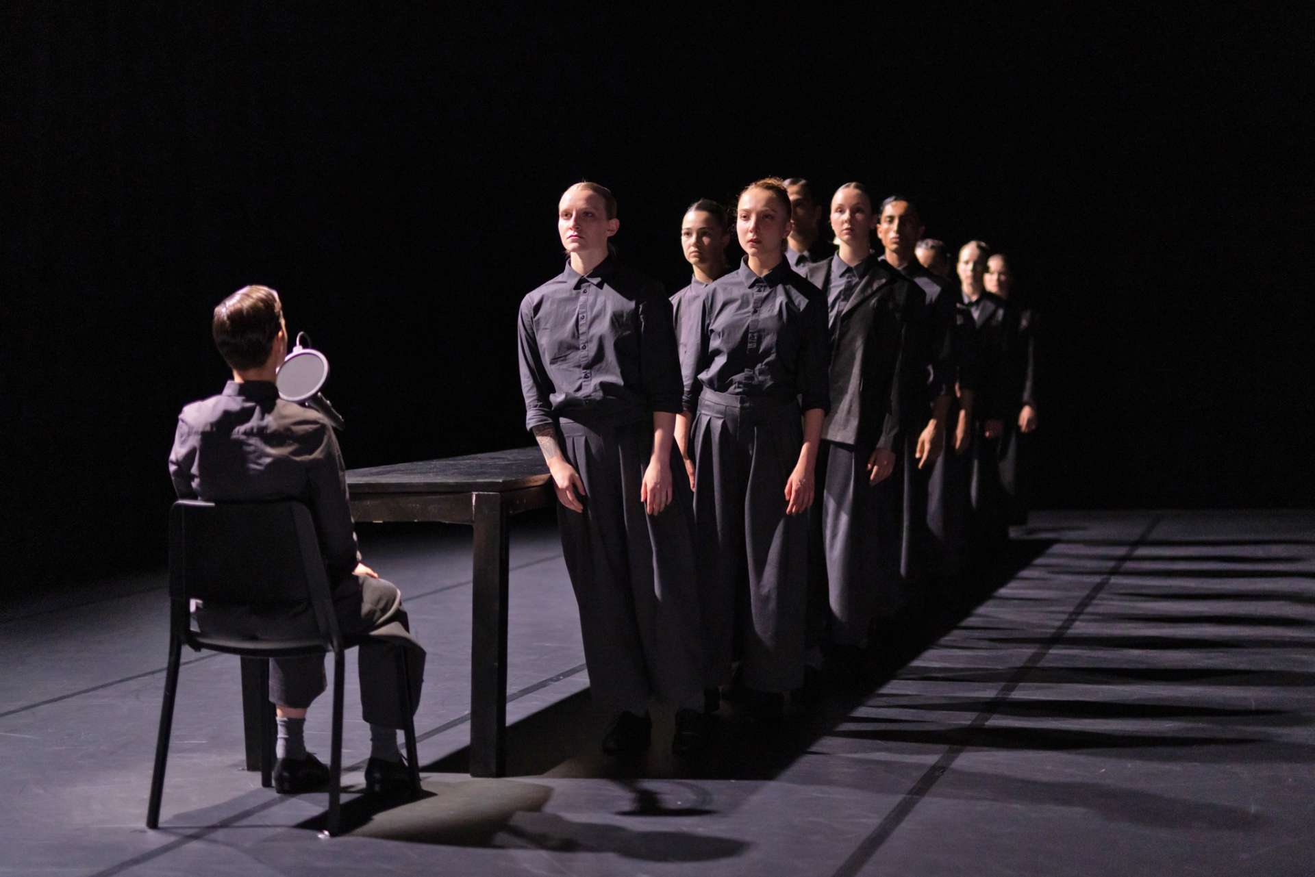 De dos, un homme est au micro. Face à lui, une ligne de danseurs et danseuses vêtus de noir et à l'air très sérieux.