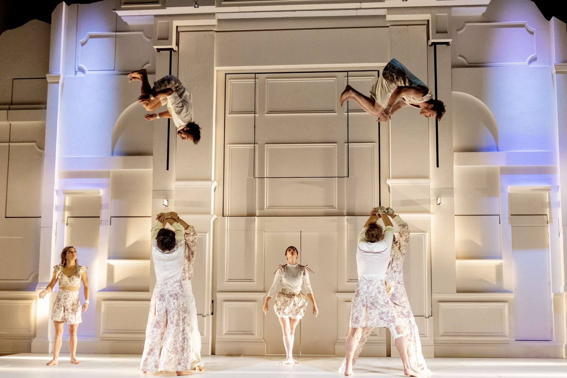 Devant un décor à moulures blanches, deux acrobates sont lancés en simultané par deux groupes de porteurs. 