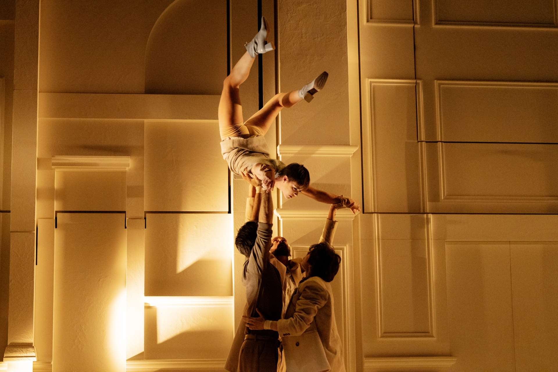 Une acrobate réalise, dans les airs, la lettre C avec son corps alors que d'autres interprètes la porte. 