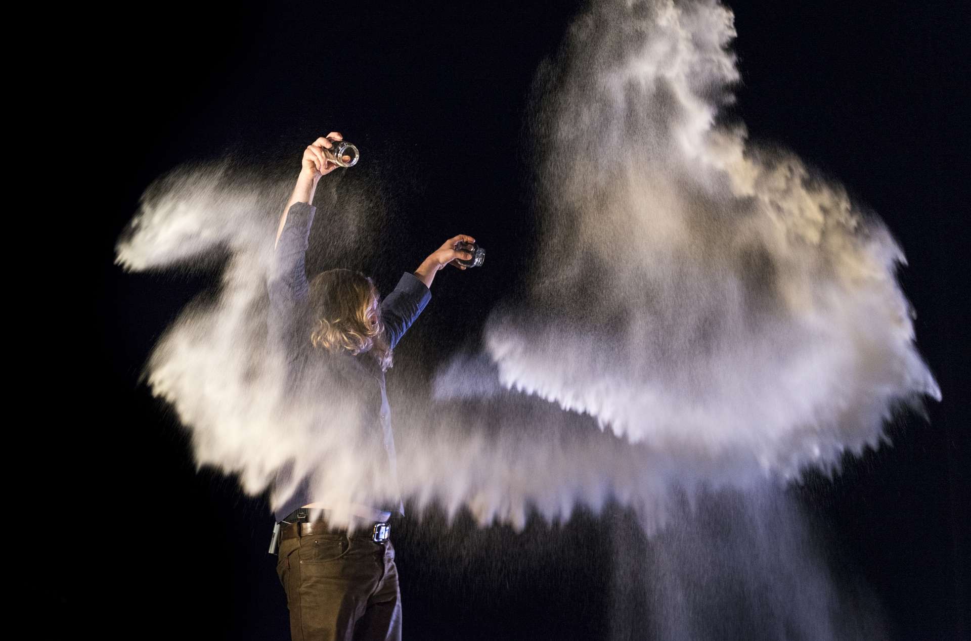 Le magicien Kurt Demey apparait sur scène les bras levés dans un nuage de poudre. La poudre est issue des pots qu'il tient dans chacune de ses mains. 