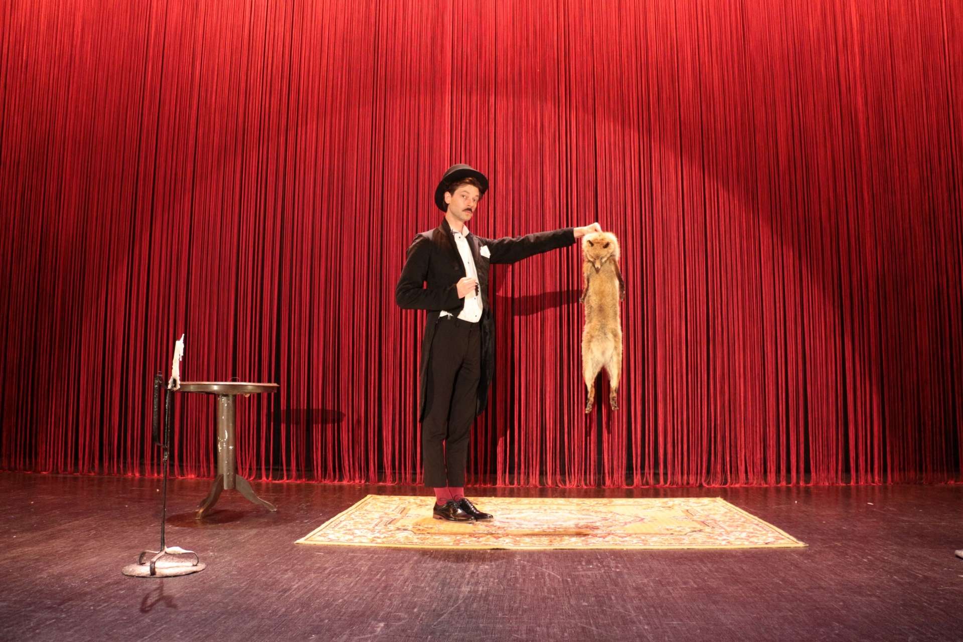 Devant un fond rouge, le grand magicien Kosmao présente son assistant Goupil. Ce dernier est un renard taxidermisé qu'il tient par la peau du cou. 