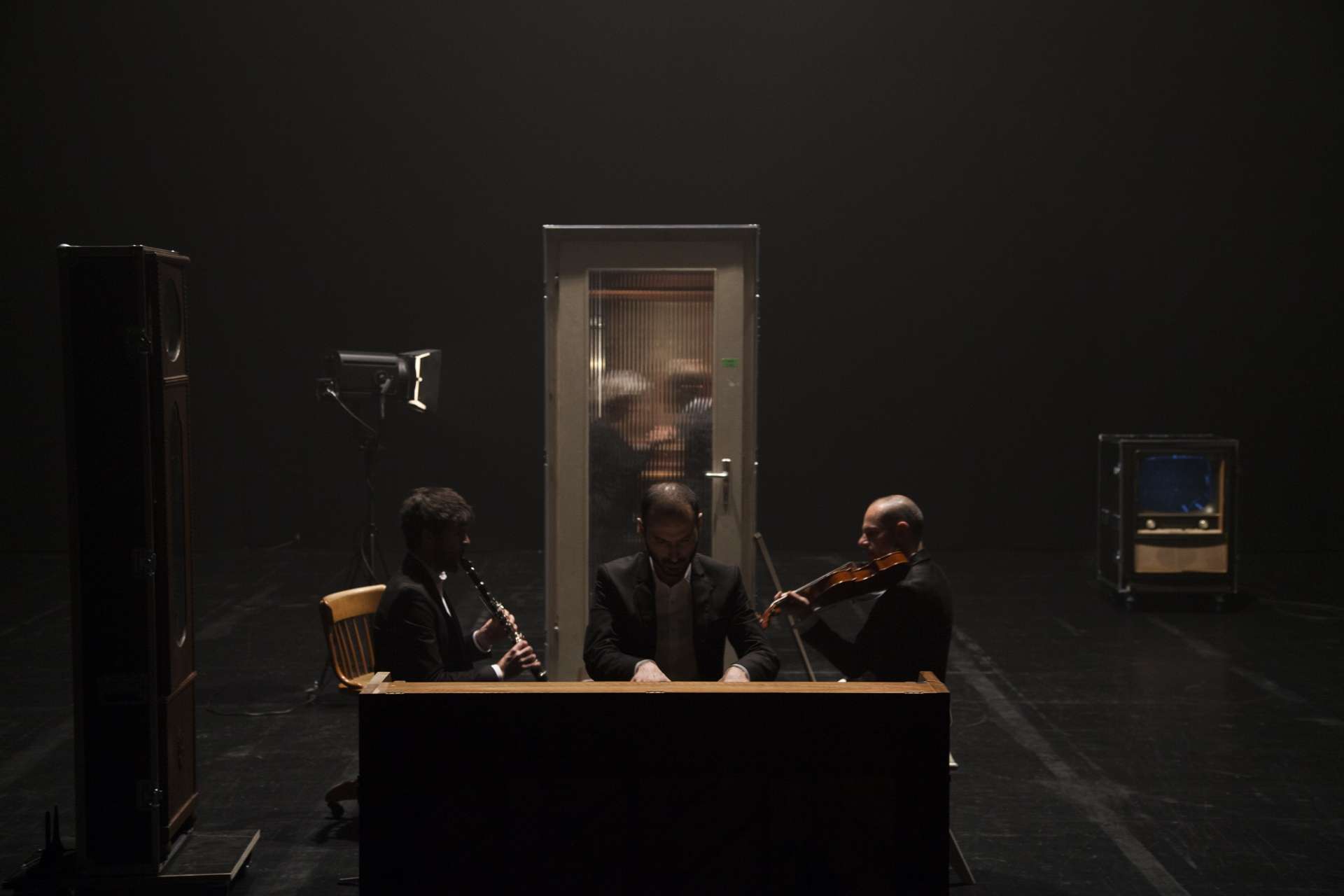 Dans la pénombre, un flûtiste et un violoniste joue autour d'un pianiste. Au fond, une cabine est éclairée. 