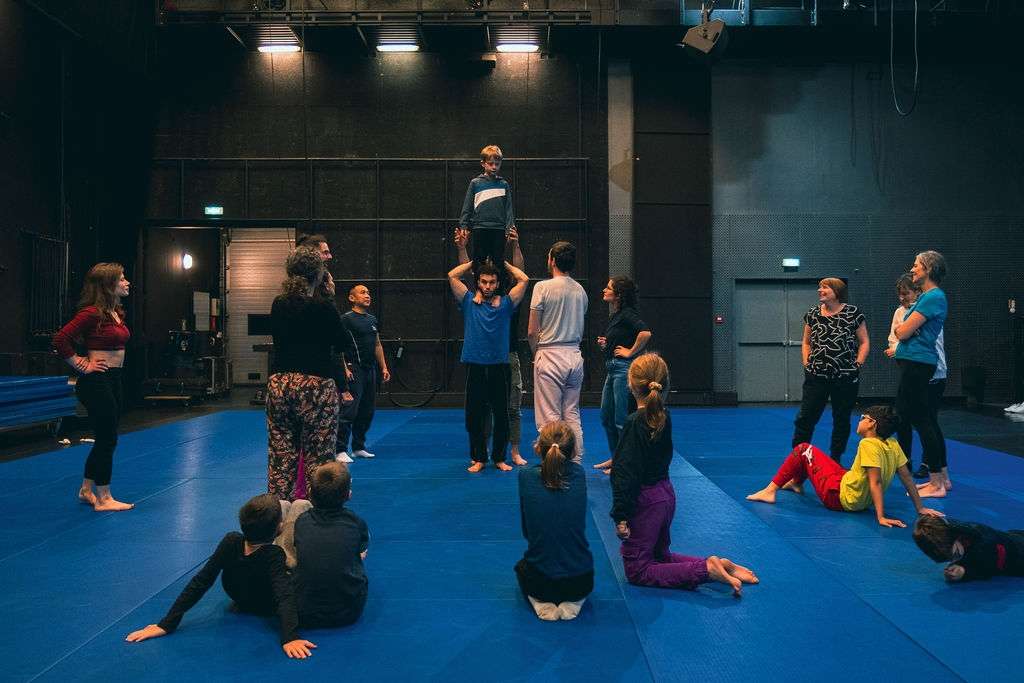 Un jeune garçon en équilibre sur les épaules d'un homme et entouré d'enfants qui les regardent, lors d'un atelier de cirque en intérieur.