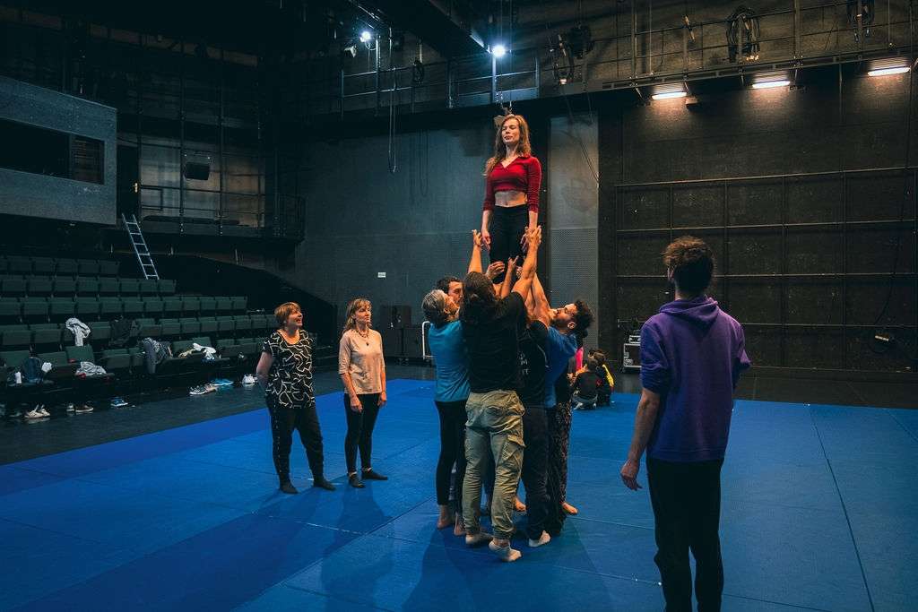 Une femme en équilibre au sommet d'une pyramide humaine, soutenue par plusieurs personnes lors d'un atelier de cirque en intérieur.