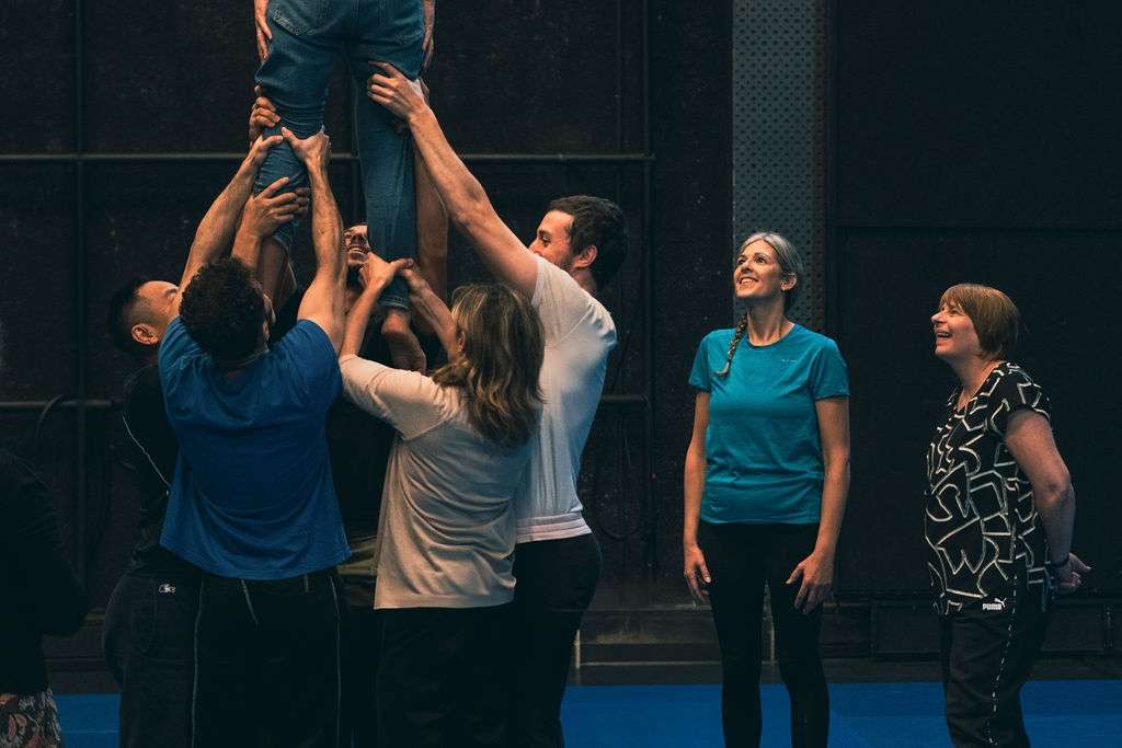 En plan rapproché, un homme de dos en équilibre au sommet d'une pyramide humaine, soutenue par cinq adultes lors d'un atelier de cirque en intérieur. Deux femmes regardent la démonstration.