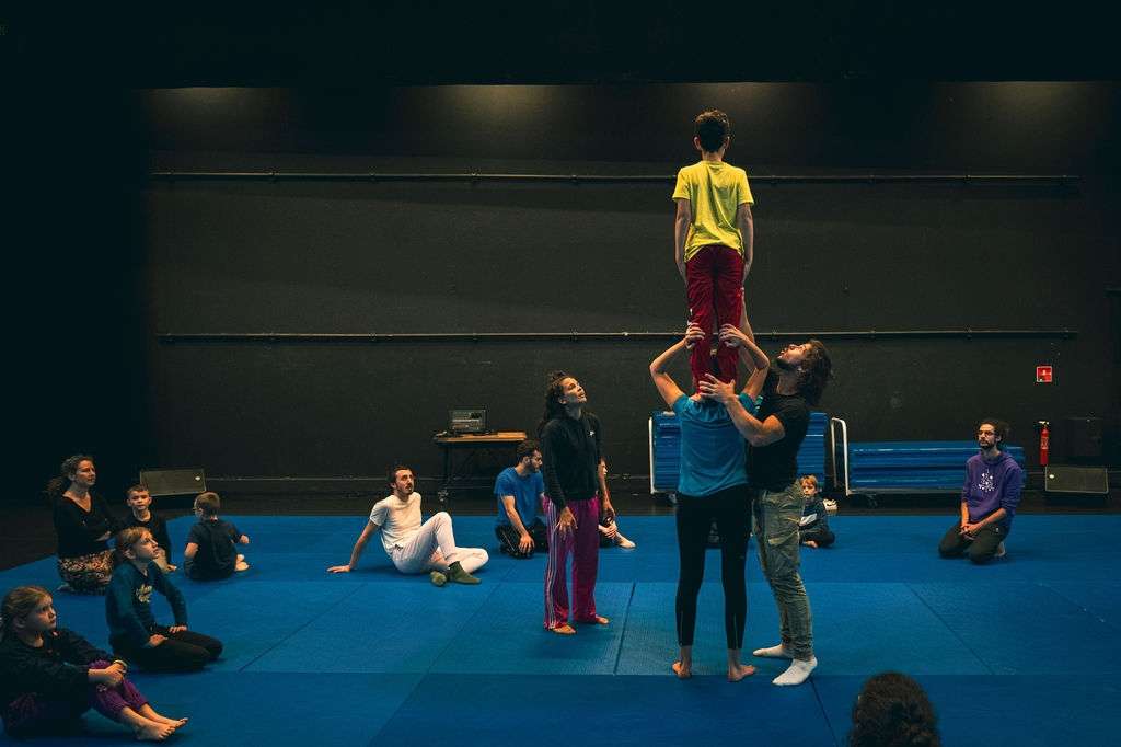 Un jeune garçon en équilibre sur les épaules d'une femme, assuré par un homme. Plusieurs personnes regardent la démonstration lors d'un atelier de cirque en intérieur.