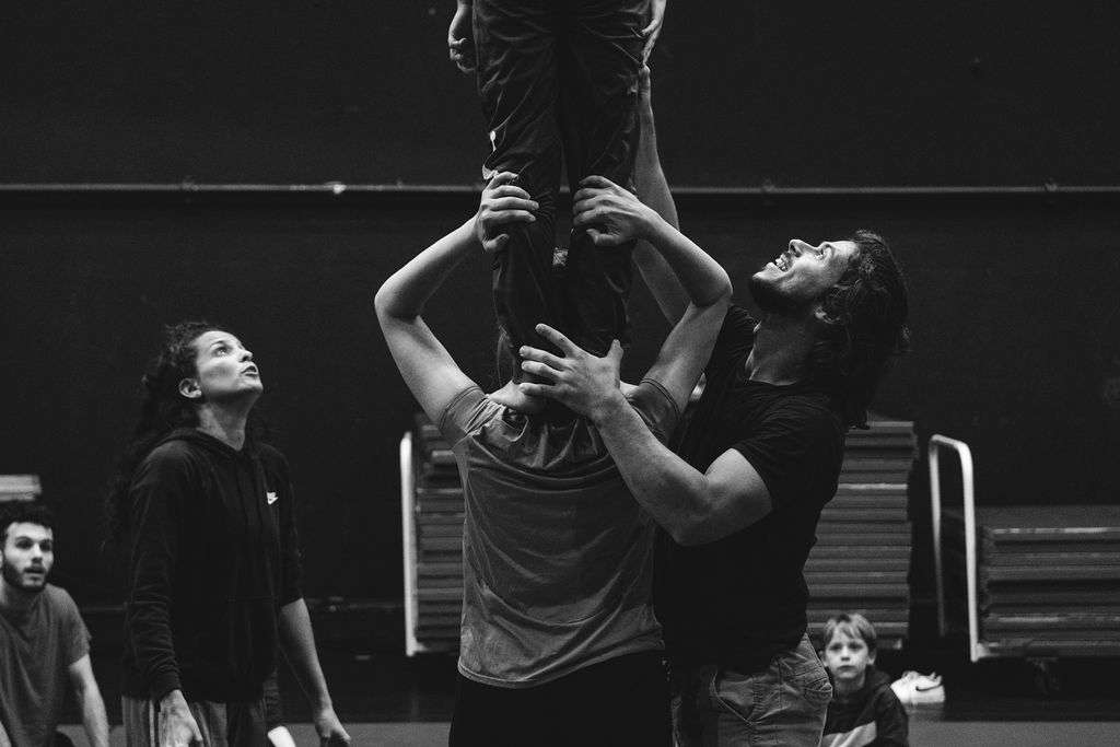 Photo monochrome en gros plan, lors d'un atelier cirque un homme tient sur ses épaules une personne. Un autre homme assure le porté et une femme regarde et donne des conseils.