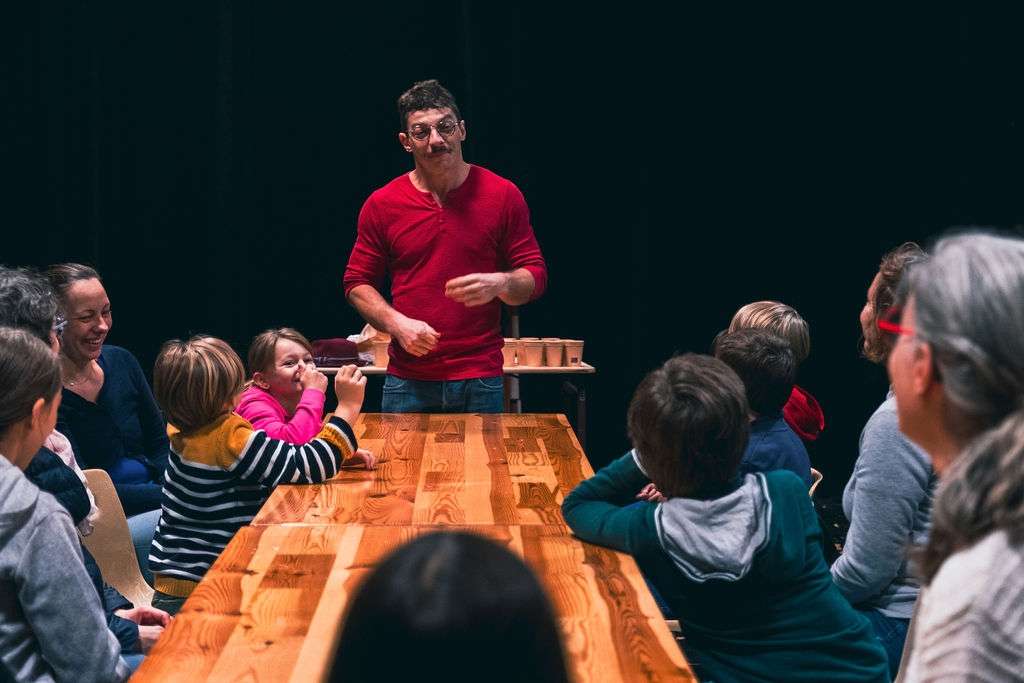 Un homme en rouge anime un atelier autour d'une table en bois, entouré d'enfants et d'adultes attentifs.
