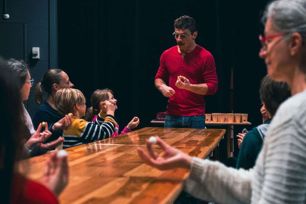 Un homme en rouge anime un atelier de magie autour d'une table en bois, entouré d'enfants et d'adultes qui participent.