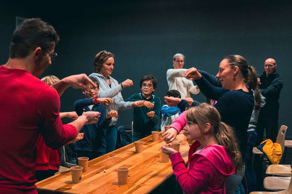 Un homme en rouge anime un atelier magie autour d'une table en bois, entouré d'enfants et d'adultes. 