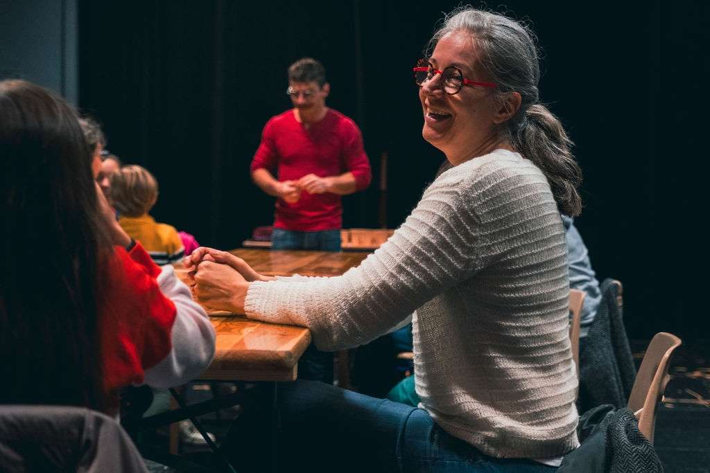 Une femme en premier plan qui sourit à une jeune fille assise à coté d'elle, lors d'un atelier cirque animé par un homme en arrière plan.