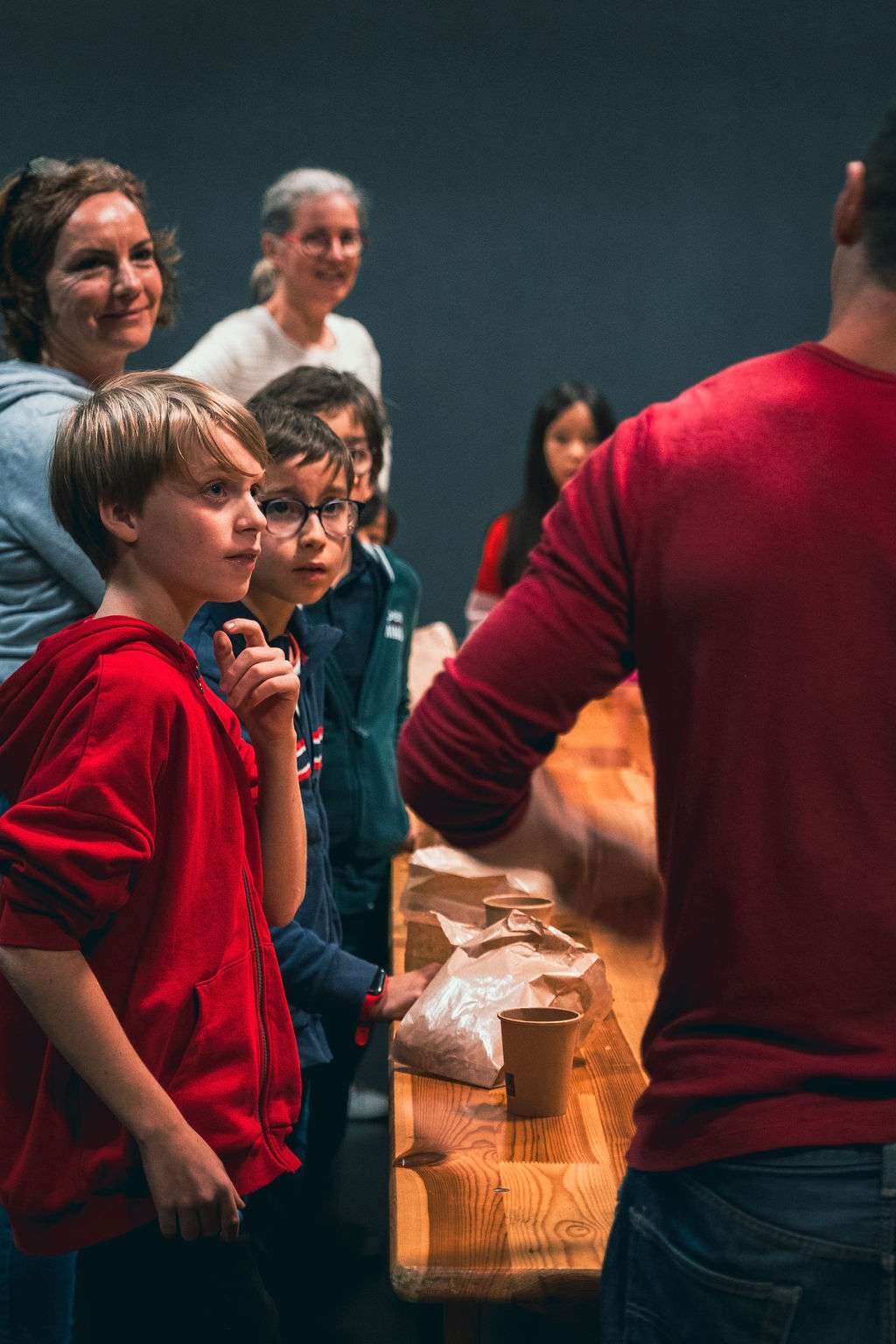 Un homme en rouge anime un atelier autour d'une table en bois, entouré d'enfants et d'adultes attentifs.