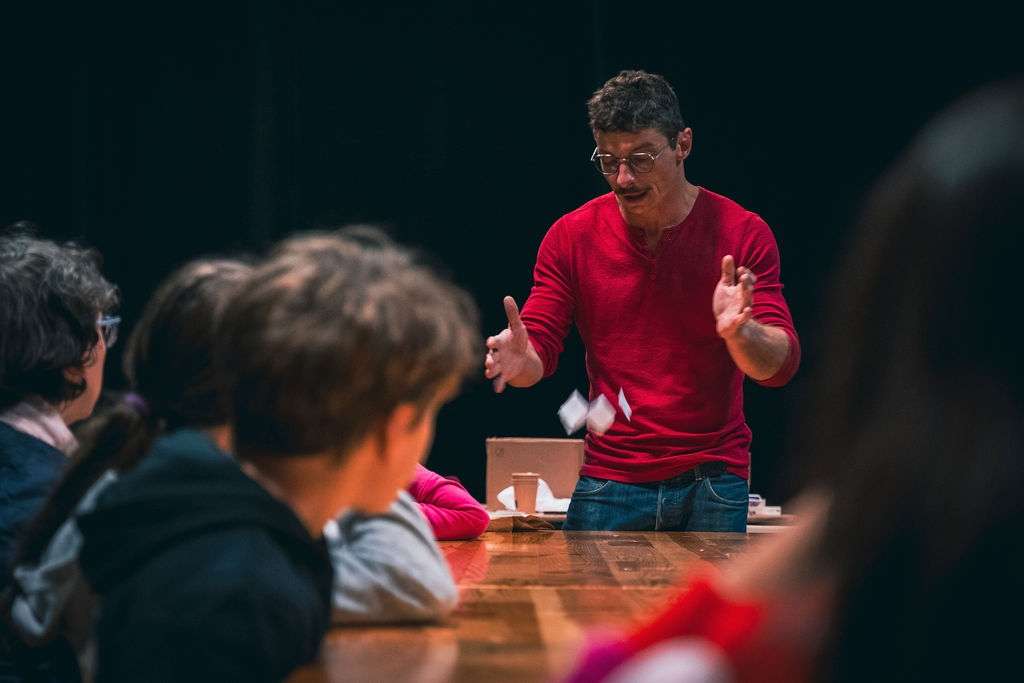 Un homme en rouge anime un atelier magie autour d'une table en bois, et montre un tour de magie avec des cartes, entouré d'enfants et d'adultes attentifs.