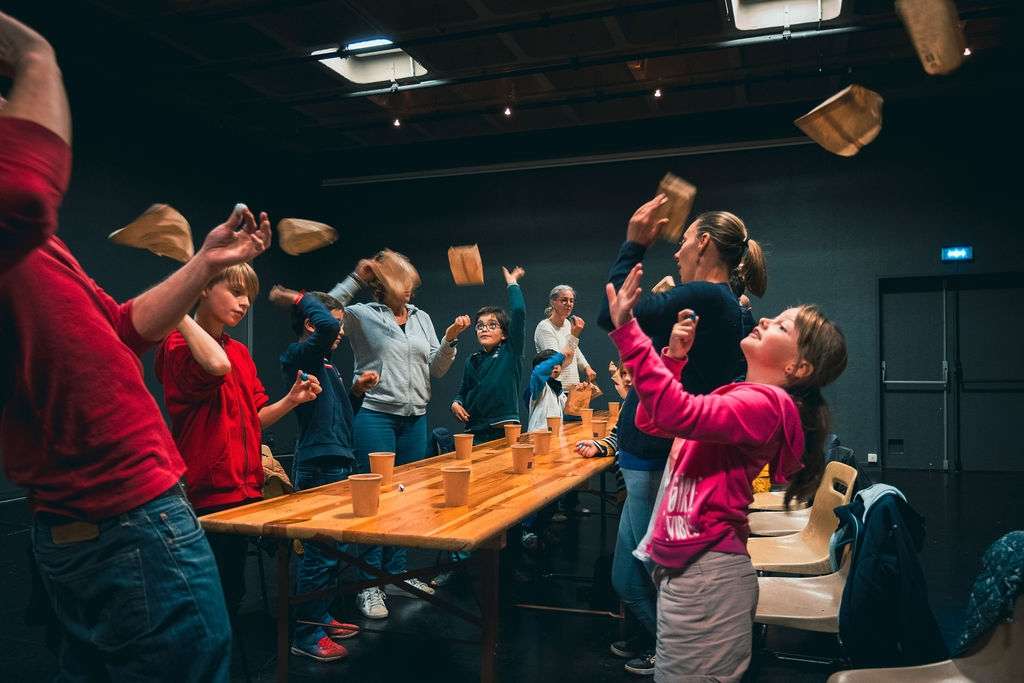 Des adultes et des enfants lors un atelier magie, autour d'une table en bois, lancent en l'air un sac kraft.