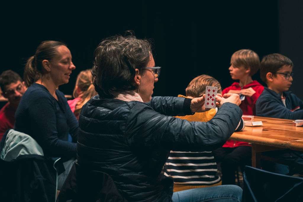 Une femme en premier plan qui teste un tour de magie avec des cartes, autour d'une table en bois, entourée d'enfants et d'adultes qui s'amusent.