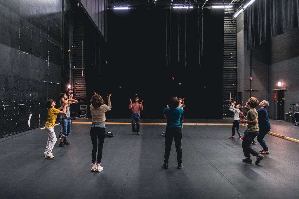 Plan large, des adultes et des enfants debout en cercle lors d'un atelier, jonglant avec des balles, concentrés sur leurs mouvements.