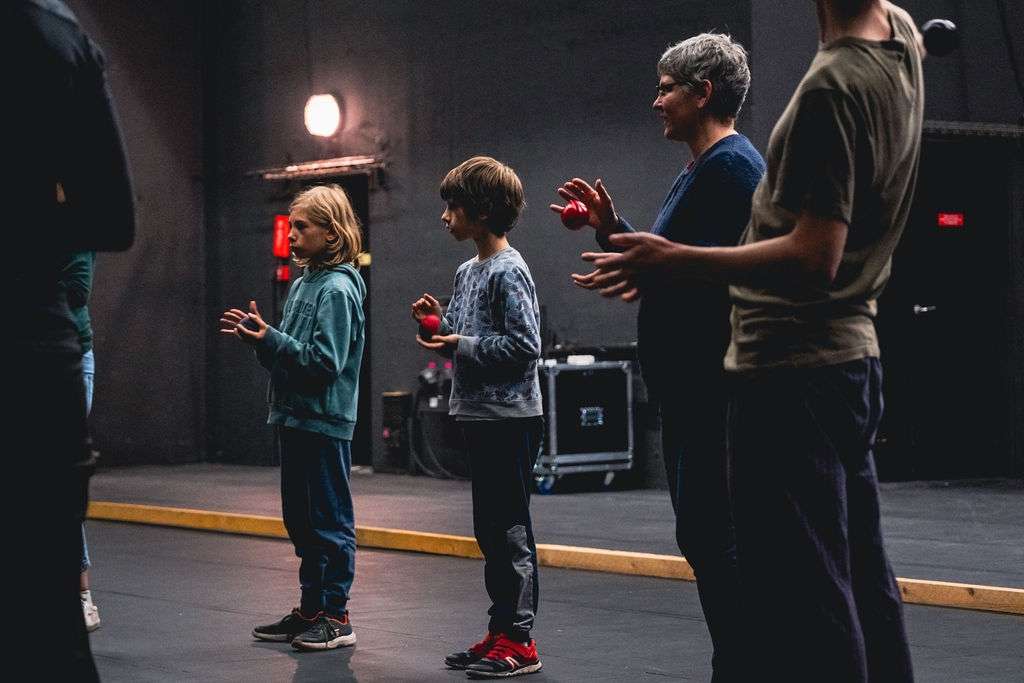 Des adultes et des enfants debout en cercle lors d'un atelier, écoutant les consignes avec des balles dans la main.
