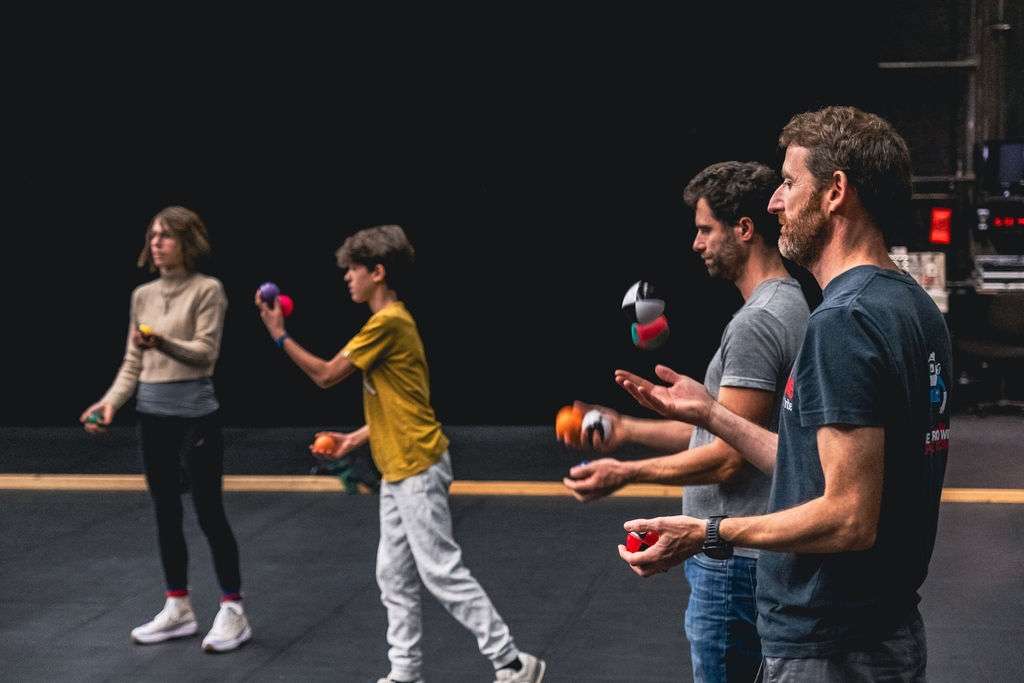 Des adultes et des enfants debout lors d'un atelier cirque, certains jonglant avec des balles, concentrés sur leurs mouvements.
