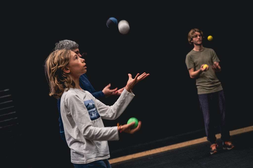 Focus sur un jeune garçon jonglant avec des balles, concentré sur ses mouvements. Deux adultes jonglant aussi derrière lui.