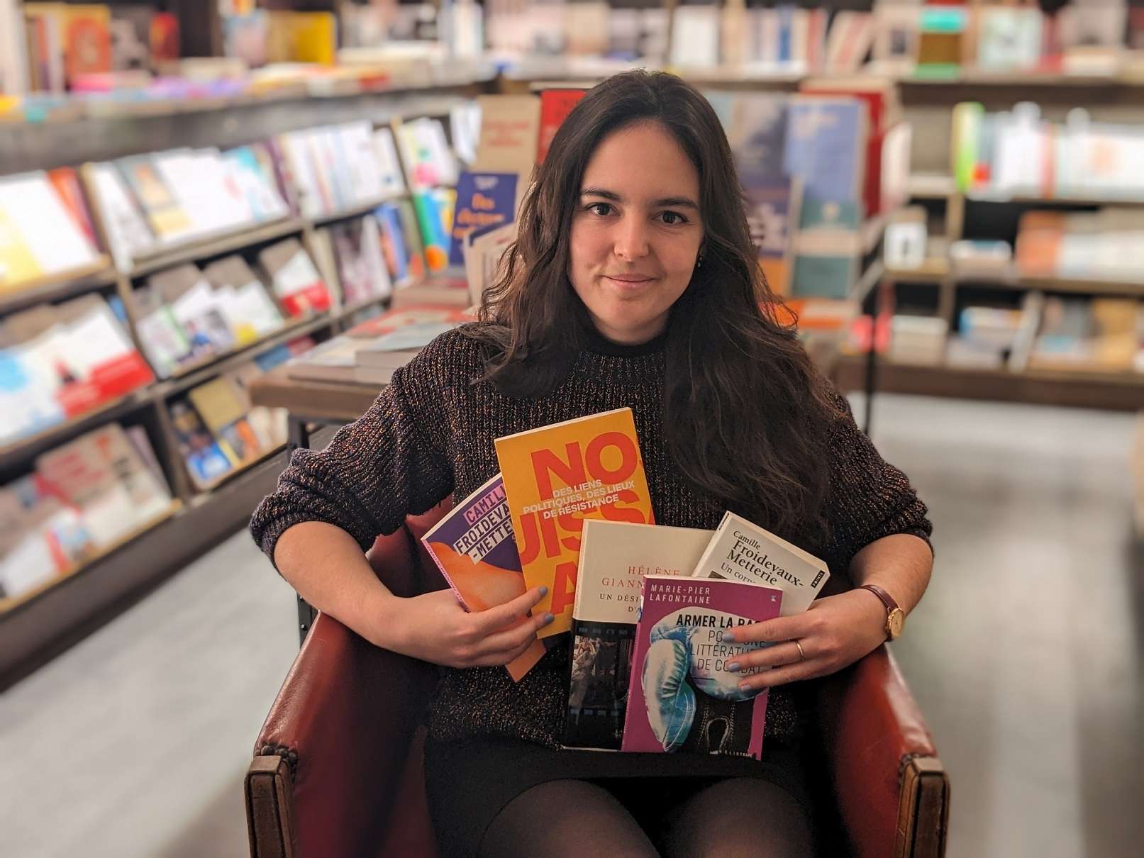 Deborah Vedel, libraire chez Les Temps Modernes à Orléans, est assise sur un fauteuil et tient des livres dans ses bras.