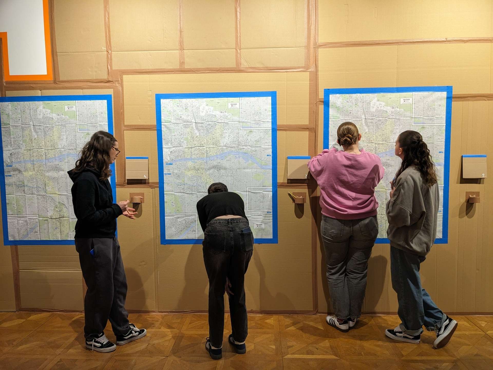 Quatre jeunes femmes sont en train d'écrire sur des cartes de la ville. Le mur sur lequel sont accrochées les cartes est recouvert de cartons.