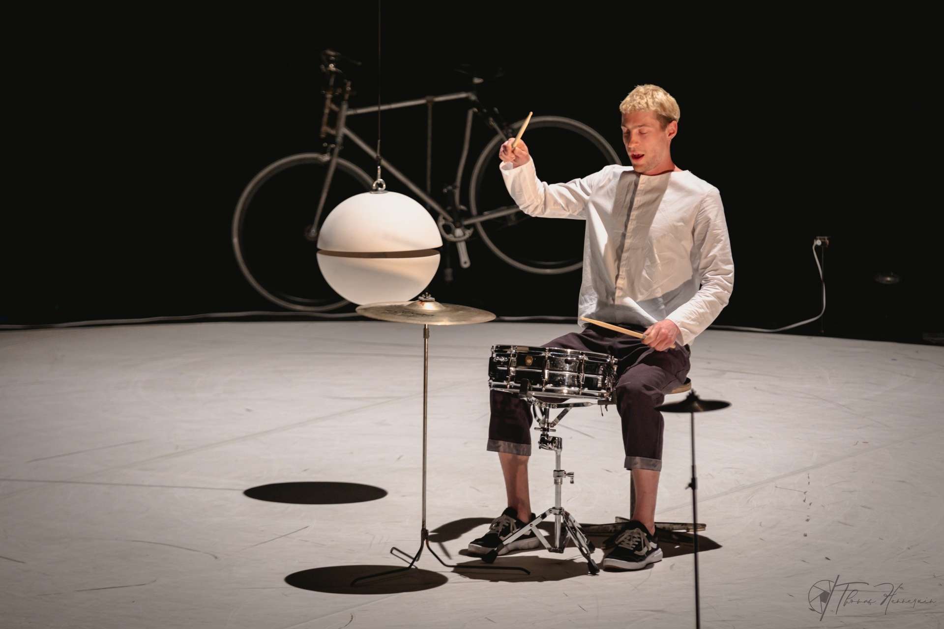Au milieu de la piste blanche, l'artiste Julian Vogel est assis et joue de la batterie. Une grosse boule est suspendue en l'air et on aperçoit un vélo en fond de scène. 
