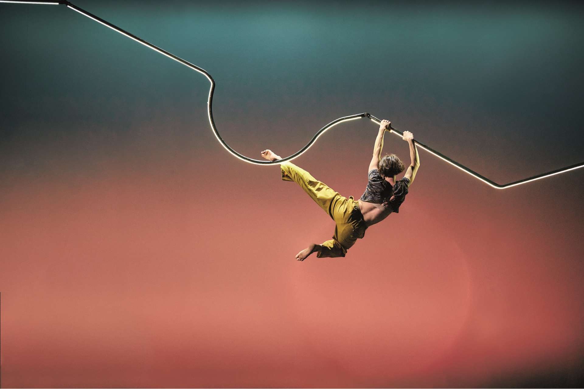Dans une lumière digne d'un beau couché de soleil, une acrobate est suspendue dans les airs, accrochée à une barre métallique qui ondule. 