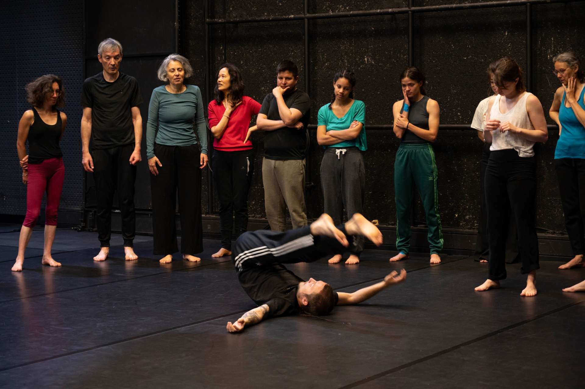 Photo de l'atelier de danse acrobatique, Clotaire en premier plan allongé sur le sol, montre une figure. Les autres participant·es suivent son mouvement et regardent.