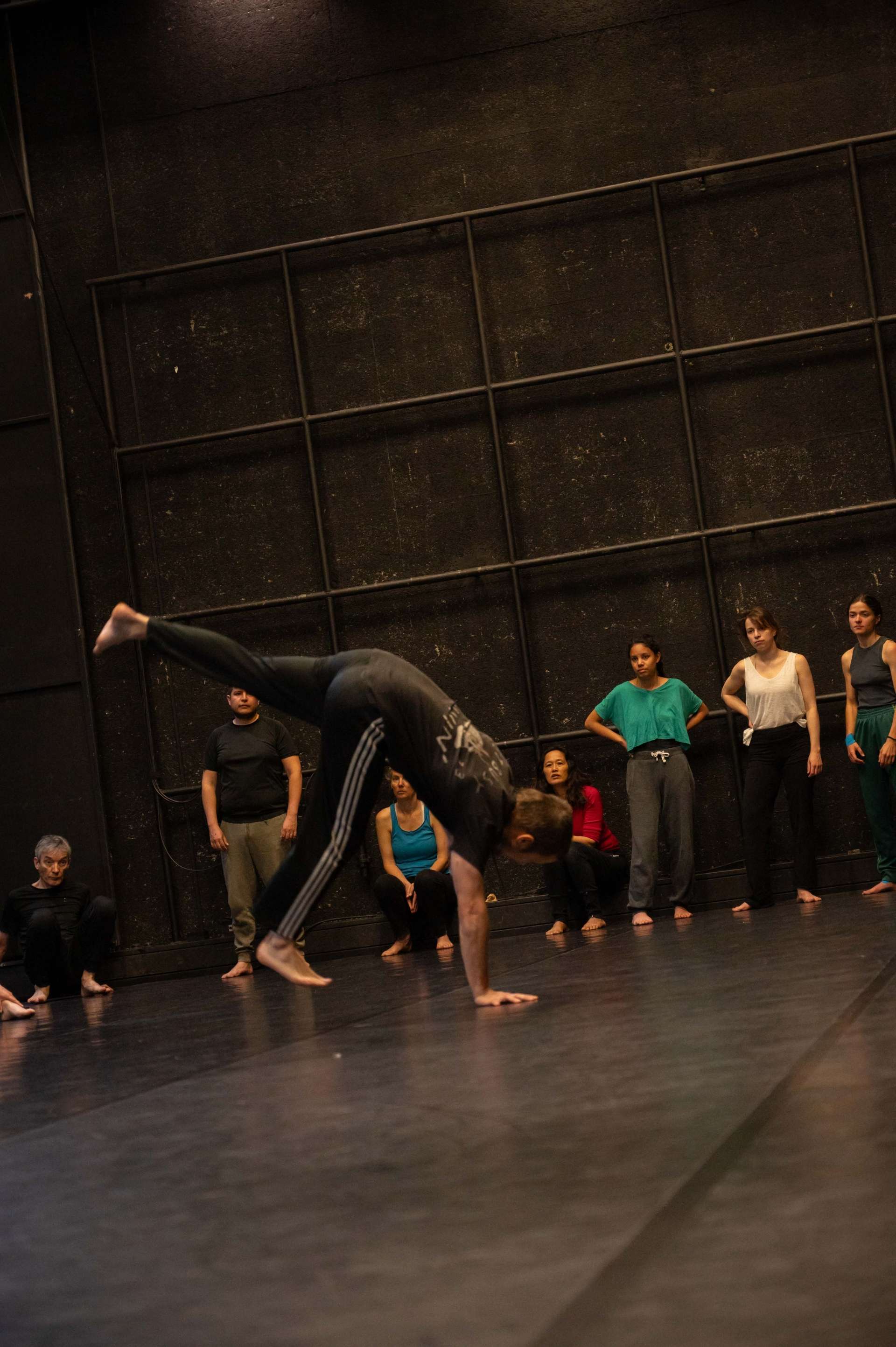 Photo de l'atelier de danse acrobatique, focus sur Clotaire qui fait un poirier, les participant·es au loin qui le regarde. 